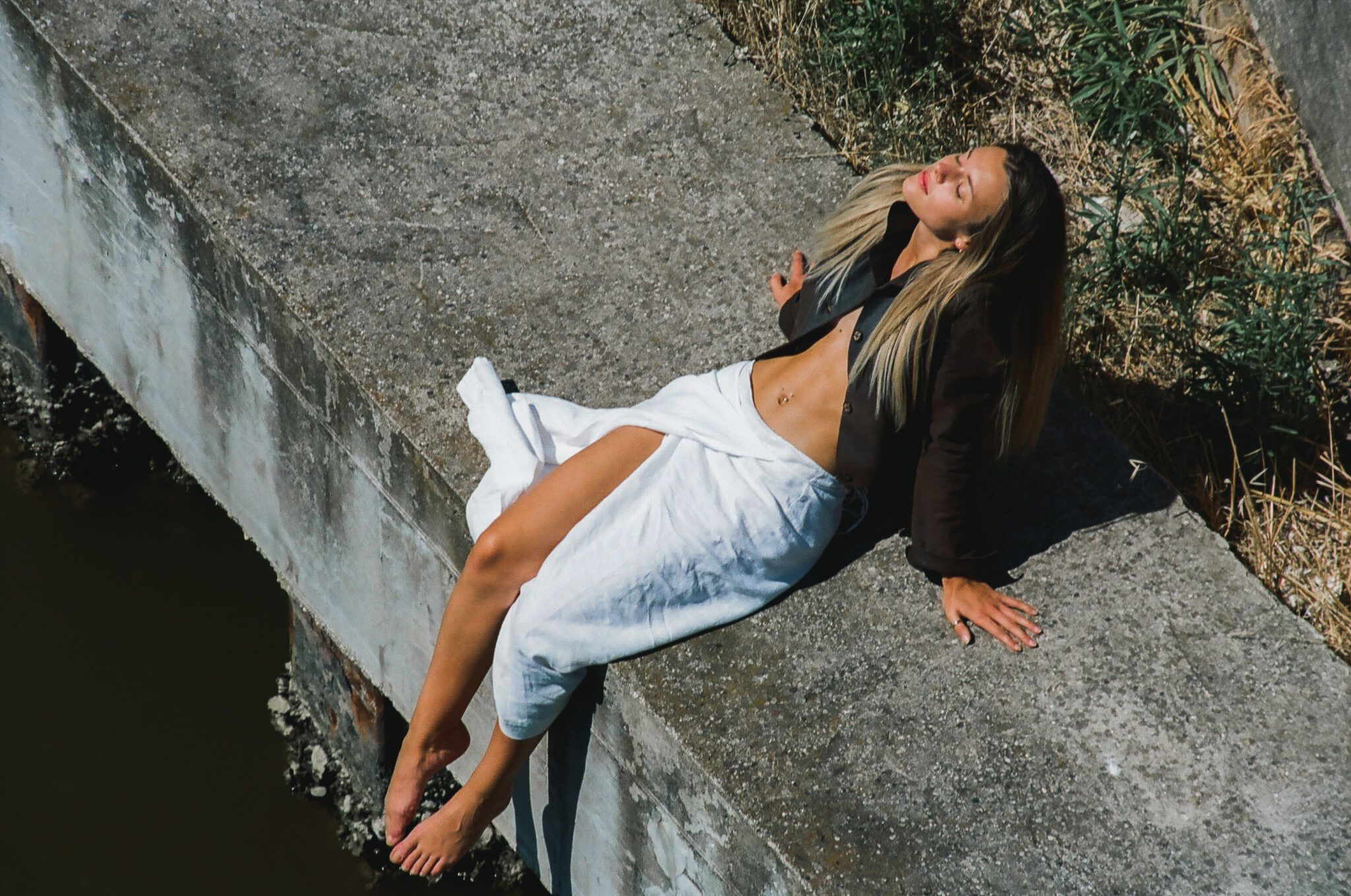 Modella bionda con gonna a portafoglio bianca, giacca di pelle marrone, prende il sole seduta su un argine, fotografia analogica