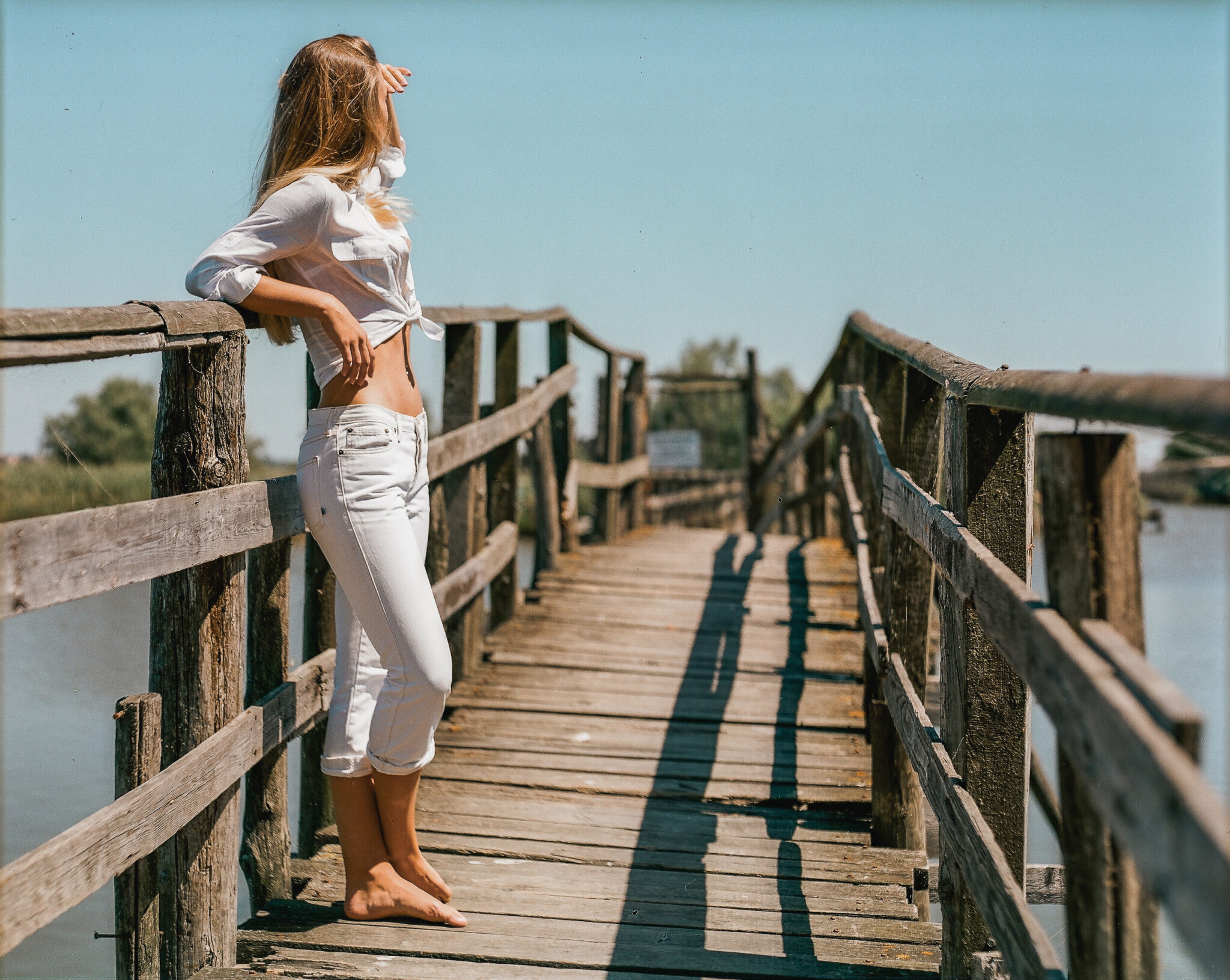 Modella con camicia bianca annodata e jeans bianchi su un ponte, guarda lontano, fotografia analogica