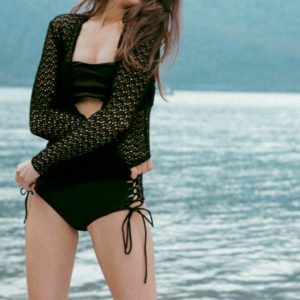 Modella con costume nero a vita alta e cardigan in macramé, con lago di Como sullo sfondo